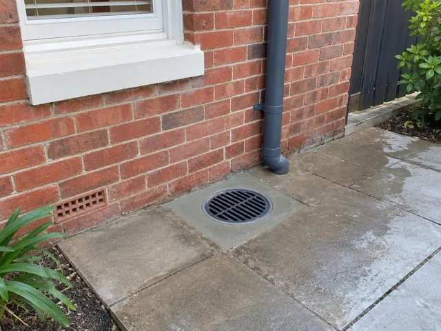 Circular outdoor drain grate set in concrete paving beside a brick wall and downpipe in Islington